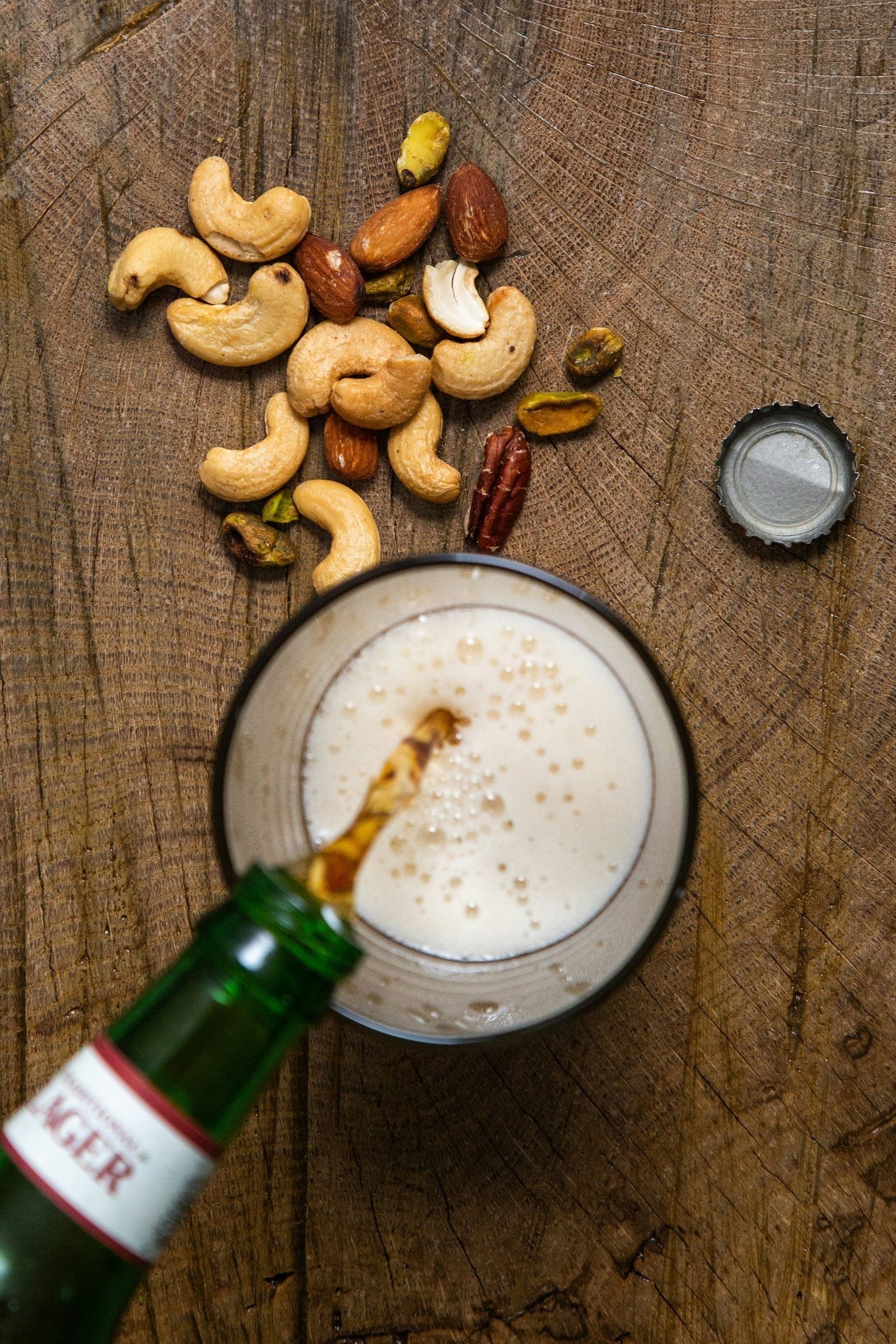 Close-up of a handful of mixed nuts scattered on a wooden table beside a glass of foamy beer, with a green-labeled lager bottle partially visible as it pours beer into the glass.