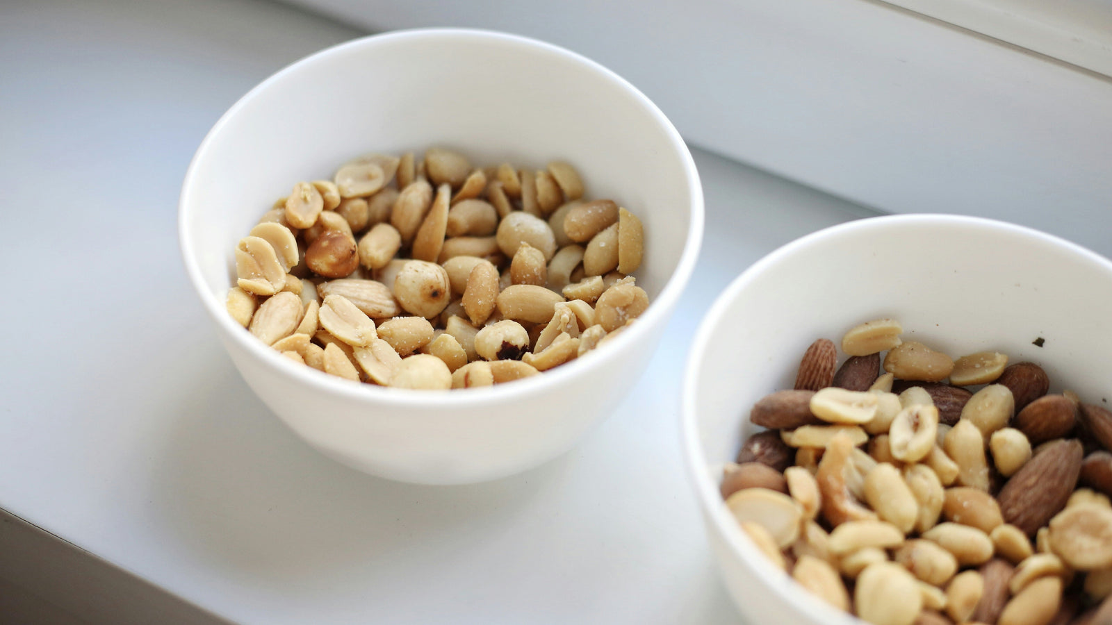 Two small, white bowls of peanuts and nuts side by side on a white table.
