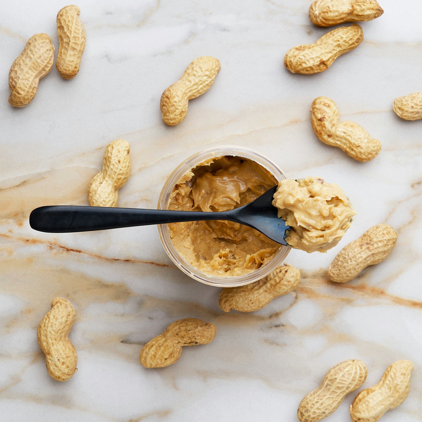 Whole in-shell peanuts scattered across a white countertop with subtle gold marble-like veining. At the center sits an open jar of peanut butter, topped with a black spoon resting across the jar’s rim, holding a scoop of peanut butter.