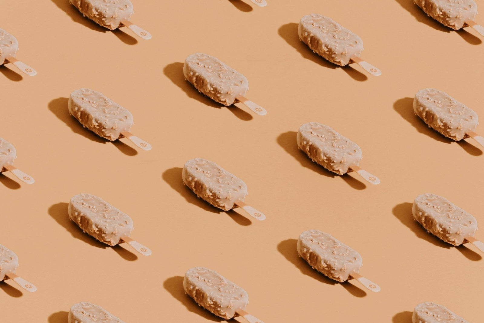 Rows of frozen peanut ice cream bars coated in chopped peanuts, arranged diagonally with space between each bar, on a light orange surface.