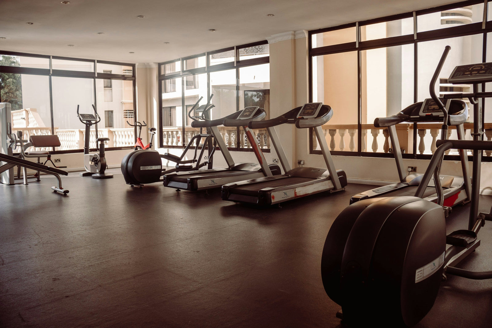 Image of an empty workout room filled with treadmills, ellipticals, and other gym equipment. A wall of windows lets in natural light.  